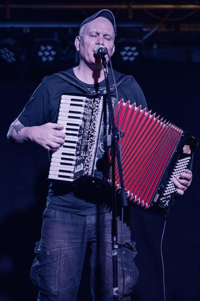 A man is performing on stage, playing an accordion and singing into a microphone. He is wearing a black t-shirt and a cap, with his eyes closed in concentration. The background is dark, highlighting the performer and the red and black accordion.