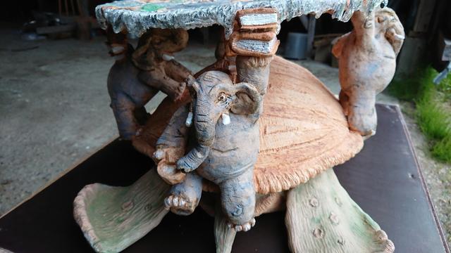 A'Tuin from the back showing one of the elephants holding two books in its hand and on top of those it's holding its part of tge Discworld. There's two more elephants behind