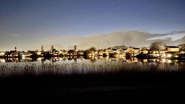 A calm body of water reflecting illuminated buildings along the shoreline at dusk. The sky transitions from twilight to night, with soft clouds present and a few stars visible. Silhouetted reeds emerge in the foreground.