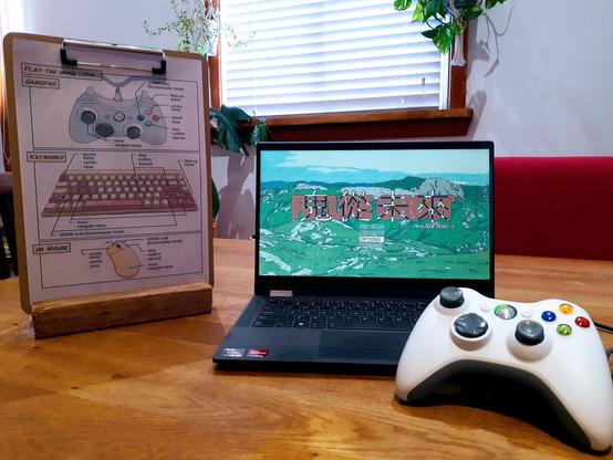 Laptop computer with the game "Future Ghost' running, an Xbox 360 controller plugged into it, sits on a timber tabletop. Next to it is a clipboard with a sheet of paper indicating the game's controls on gamepad, keyboard and mouse. The clipboard stands vertically, held up by a stand fashioned from a wooden block.