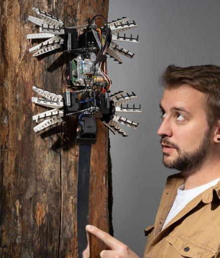 A photo of Rob next to a lizard-inspired robot on a tree. He's wearing a tan shirt over a white t-shirt.