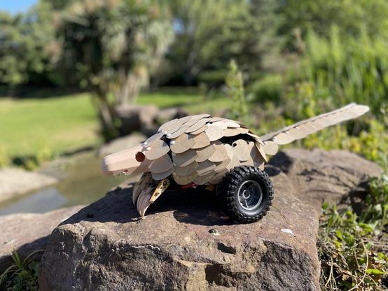 A beige scaley robot with a long snout, two clawed front paws for digging, a long scaley tail and two large black wheels. It's sitting on a rock.