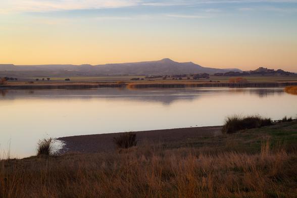 Fotografía en la que se puede apreciar un paisaje al atardecer con unas montañas bajas al fondo, una capa de agua en el medio y algo de hierba y ramas secas en la orilla en primer plano. Todo ello con una suave luz del atardecer. | Photography in which you can see a evening landscape with some low mountains at background, a layer of water in the middle and some grass and dry branches at the shore in the foreground. Everything with a smooth light of evening.