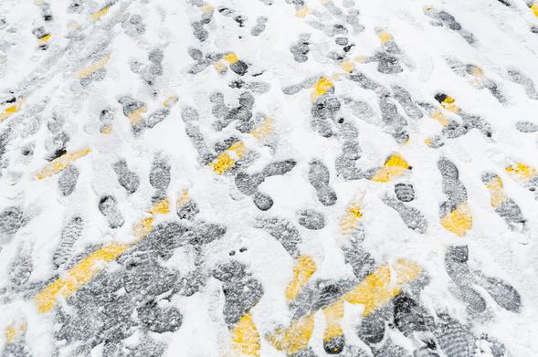 Footsteps in snow reveal the carpark and its markings beneath. Bright yellow diagonal lines juxtaposed with the grey asphalt and the bright white of snow.