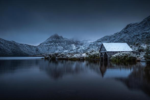 A moody early morning scene showing a dusting of snow at Dove Lake in Tasmania. On the right is a boat shed reflected in the still waters with snow on its roof. On the left is lake and the mountain itself, partially obscured by the low snow clouds.