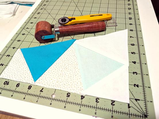 Half a quilt block, if the square of it were cut at a 60-degree angle down the middle.  Triangles in medium blue, light blue, white with gold metallic spots, and white (plain) are pointed at the center.  The corners are white & white with gold metallic spots.  It rests on a cutting mat, next to a wooden seam roller, a rotary cutter, and a clear quilter's 6" ruler.