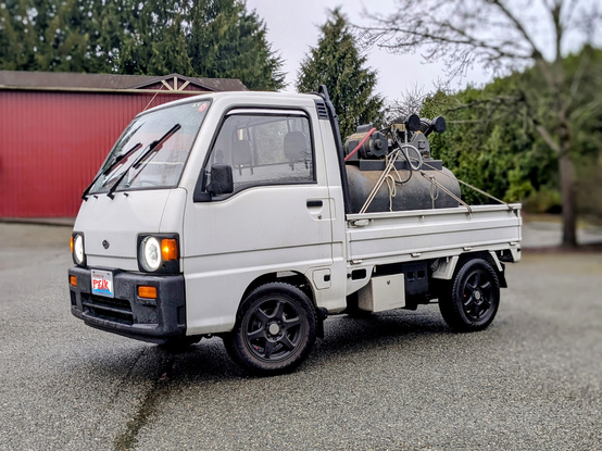 A large, dirty air compressor in the back of a white 1992 Subaru Sambar "Kei Truck".