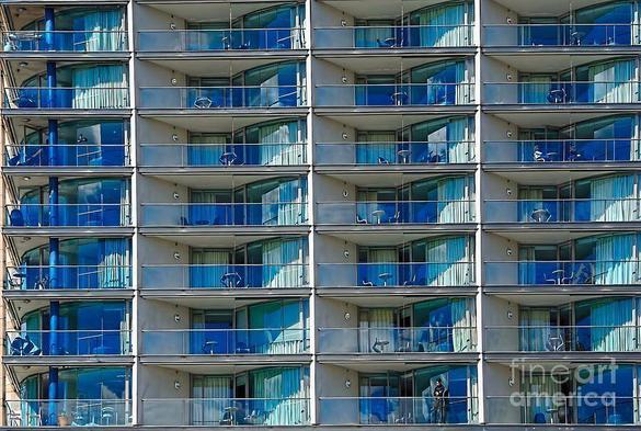I captured this image on the Eastern side of Circular Quay, Sydney, New South Wales, Australia whilst awaiting a Ferry.
I wanted to capture the repetitive similarity and pattern of the apartments.

A building facade showcases multiple floors of identical balconies with blue-tinted glass railings, creating a repetitive architectural pattern. The reflections of clouds and sky in the glass add a dynamic visual element to the structure. By Kaye Menner