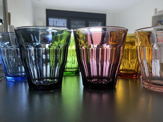 Close up picture of Duralex Picardie glasses in different colors, on a grey surface with light coming from behind the glasses, their reflections can be seen on the surface they’re on.