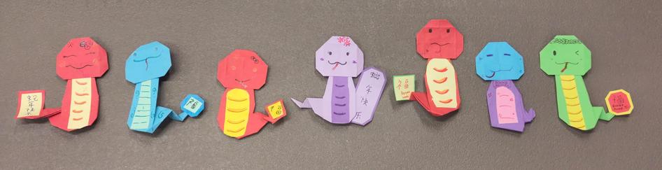 7 serpents en origami coloré, de gauche à droite : rouge, bleu, rouge, rose, rouge, mauve avec une tête bleue, et vert. 

Chaque serpent porte un petit charme sur sa queue pour souhaiter du bonheur pour la nouvelle année lunaire.