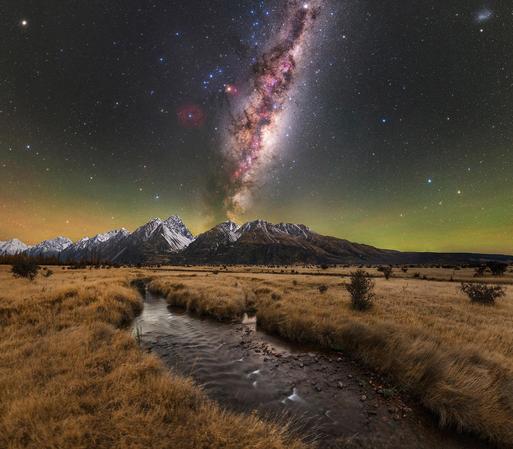 the Milky Way core, rising above a small river in New Zealand. in the background, the snow capped southern alps mountains can be seen