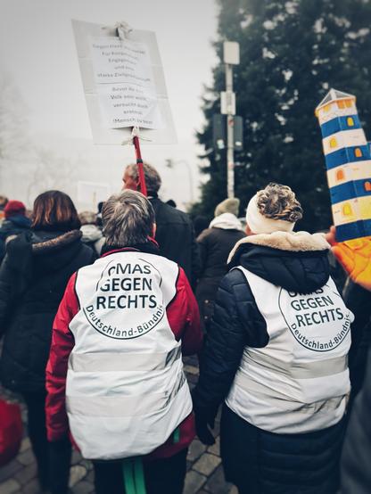 Two women participate in a protest, wearing white vests labeled "OMAS GEGEN RECHTS" (Grandmothers Against the Right). One holds a sign with messages against hate and for social engagement. The scene is foggy.
