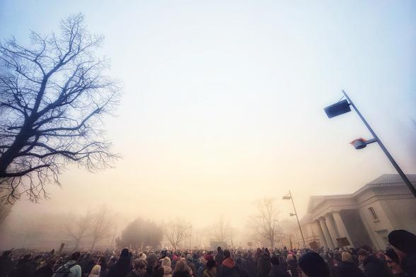 A dense crowd gathered in a foggy outdoor space, with bare trees and streetlights visible. The background features a building partially obscured by mist, creating a somber atmosphere.