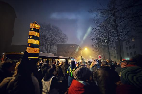 A crowded night scene at a protest or celebration, featuring a tall, illuminated lantern resembling a lighthouse. The atmosphere is foggy, with people holding flags and signs while streetlights cast a warm glow. Trees and buildings are visible in the background.