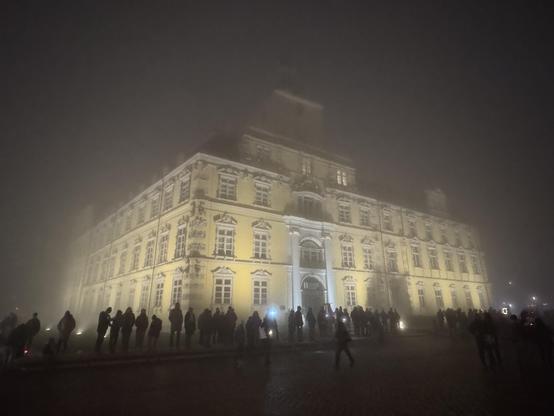 The historic yellow Oldenburg Schloss building is shrouded in fog, illuminated by soft lights in the evening. A crowd is gathered in front, creating a mysterious atmosphere.