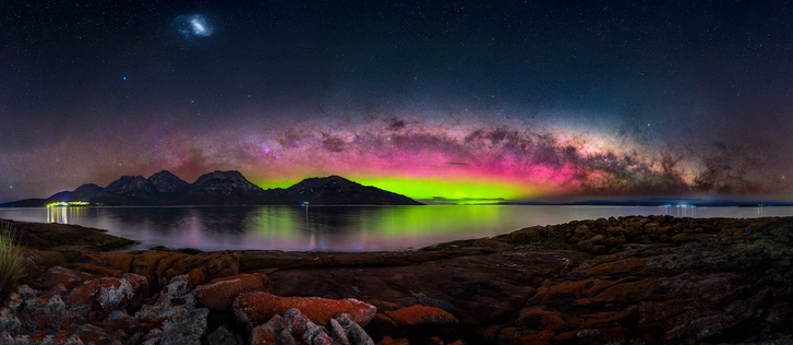 The scene is set on a wide, rocky bay with deeply red toned rocks covered in lichen. Across the bay is a jagged mountain range which rise from the edge of the bay. They have scarred dolerite faces. Behind the range is a strongly green and pink aurora, topped by the setting Milky Way over the far coastline of Tasmania’s east coast.
