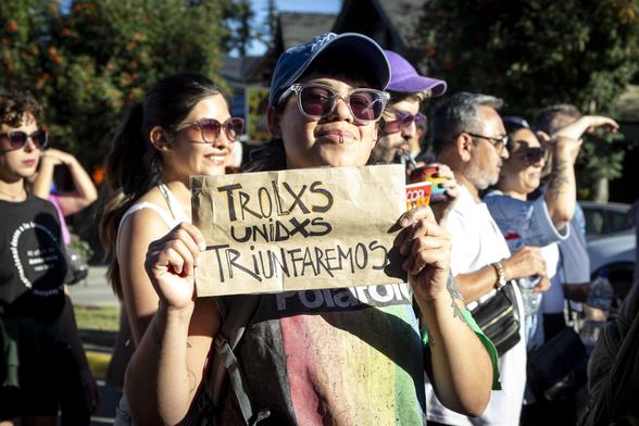 Una mujer de la comunidad lgtbiq con una remera negra con un cuadrado cuyo interior tiene rayas verticales de colores rojo, naranja, amarillo y verde; sostiene un cartel que dice en español: "Trolxs Unidos Triunfaremos". Esboza una sonrisa y su mirada parece ser de afirmación y satisfacción. Detrás de ella hay otras mujeres y hombres marchando. El clima parece ser tranquilo y festivo.