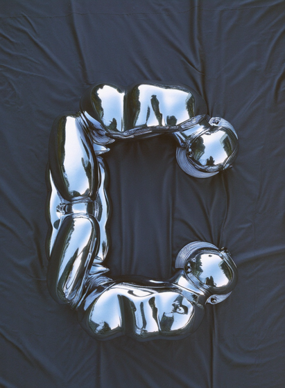  A silver, robotic-looking humanoid figure contorted into the shape of the letter "C," lying on a black fabric backdrop.