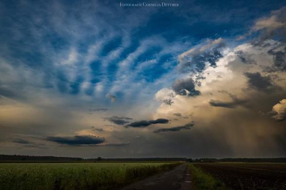 Dunkle Wolken,  links bis zur Mitte blau mit Horizontalen weißen Streifen,  rechts sieht es aus wie ein Gesicht unten im Bild ein Weg zwischen Feldern