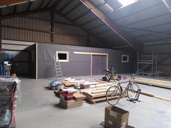 Dark grey insulation wrapped building in a hall. A few piles of wood are on the concrete floor in front of it.
