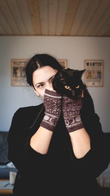 A woman wearing knitted gloves is holding a black cat in front of her. Only her eyes are visible.