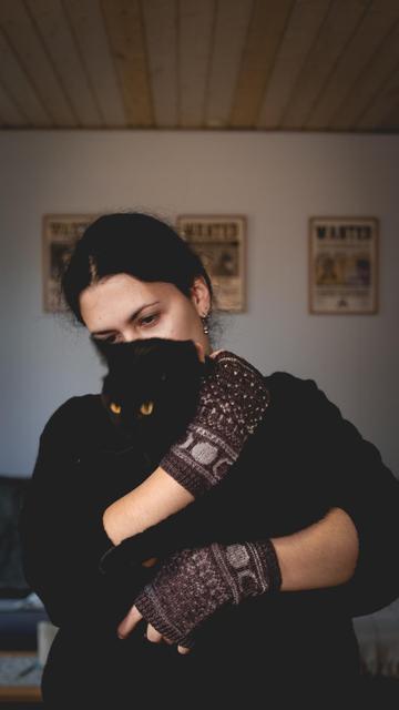 A woman wearing knitted gloves is holding a black cat in front of her. Only her eyes are visible.