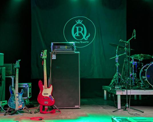 photo of an empty stage with a drumset, bass & guitar. rolling quartz logo on the black backdrop.