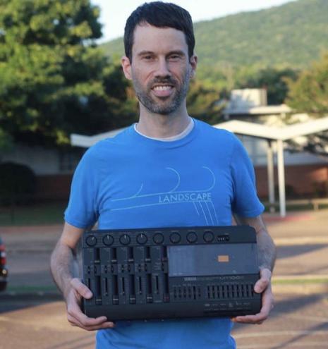 Jack Saturn stands smiling in the center of the frame, wearing a blue Landscape t-shirt while holding a Yamaha MT100 multitrack cassette recorder. There are trees and a school building behind him.