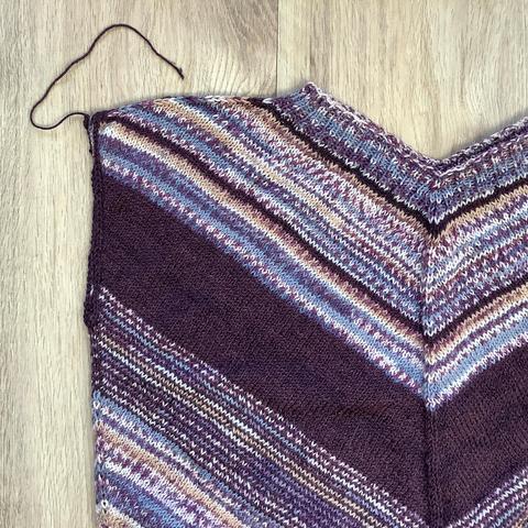 Flat lay view of a knit tee in purples, with a striking “V” shape. The armhole pictured, is bound off in aubergine colored yarn.