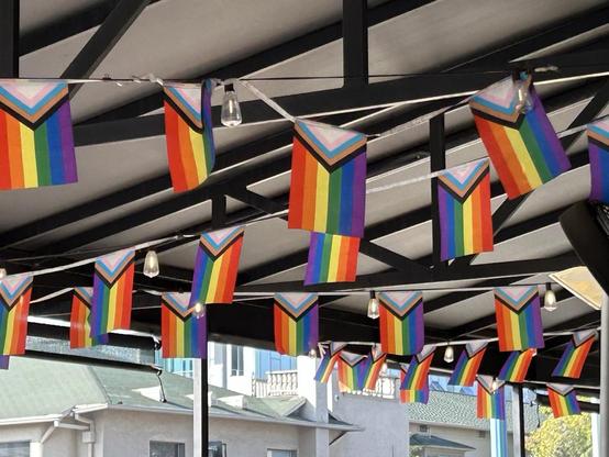 strings of progress pride flags hung up with lights under the roof of a restaurant patio 