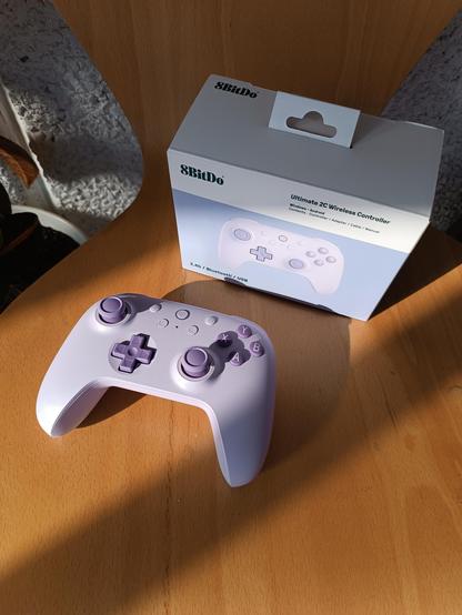 En la imagen se ve la caja de un mando de color morado junto al mando que contenía (de color lavanda y los botones de color morado más intenso). El manto tiene un formato y disposición de los botones similares a los mandos de Xbox. Ambos objetos se encuentran sobre una superficie de madera de color anaranjado.