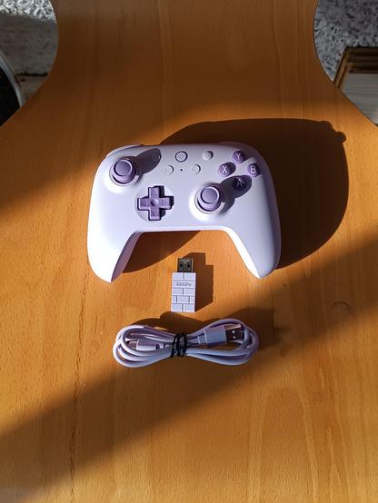 En esta imagen se ve la misma superficie de madera sobre la que reposa el mando anteriormente descrito junto con un USB que sirve para conectarlo al ordenador y un cable tipo C. Ambos, el USB y el cable, del mismo tono la banda que tiene el mando.