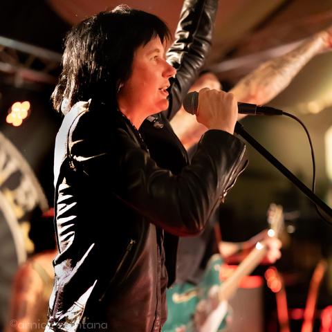 A singer in a black leather jacket passionately performs on stage, microphone in hand, with an energetic pose. The background features a dimly lit venue with blurred lights and a band member visible holding a guitar.