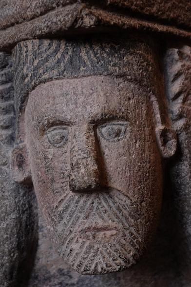 A carving in stone of a long thin face with moustache and beard, a large nose and rather round ears. It is slightly at an angle, as if quizzical. The head supports the rim of a stone font.