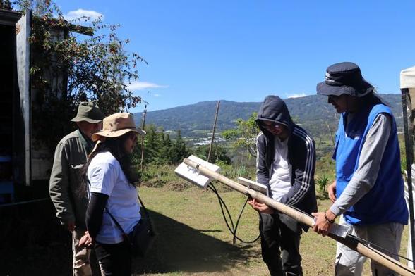 A group of four people in an outdoor rural setting examine a handmade device, possibly related to a community network project. One person in a blue vest and sun hat holds the device while explaining something. The background features green hills, a blue sky, and a small building.

Credit: A new community network, the "Red Ovejas Verdes," in Colombia. Photo: Colnodo

Source: APC Community Networks Newsletter