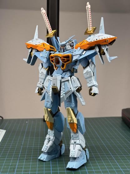 A detailed model of a robot figurine, predominantly blue with orange accents and golden parts. The figure features intricate design elements, including dual swords on its shoulders and mechanical details, posed in a standing position on a grid-patterned surface.