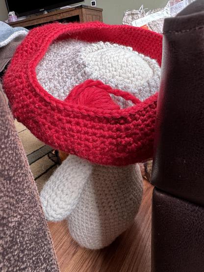 A cream crocheted plush toy is wearing an unfinished large red hat. A small ball of red yarn can be seen sitting in the hat.   The toy is positioned between two furniture pieces with a cluttered background. 