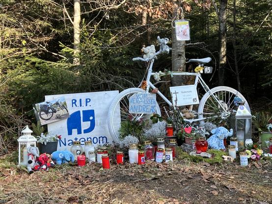 Die Gedenkstätte mit dem weißen Ghostbike. Davor stehen zahlreiche Blumen, Kerzen, Plüschelefanten und andere Plüschtiere. Am Ghostbike ist ein weißes Schild mit einem schwarzen Kreuz darauf und dem Text:

"Natenom
43 Jahre
30.01.2024"

Außerdem am Vorderrad ein Schild "Wir vermissen Andreas" und ein Transparent mit Natenoms Elefantenlogo und dem Text "RIP Natenom"