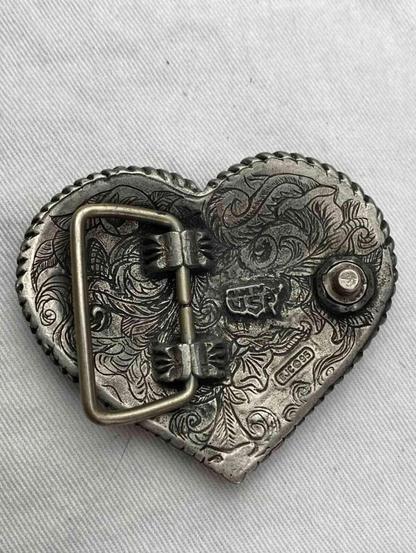 Back of a heart-shaped, ornate belt buckle with engraved floral designs and metal accents.