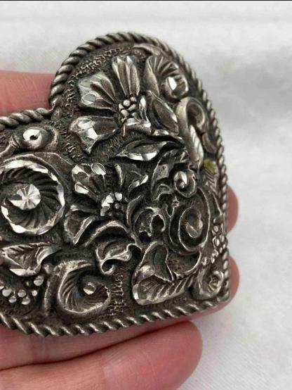 Heart-shaped, ornate belt buckle with detailed floral and vine engravings.