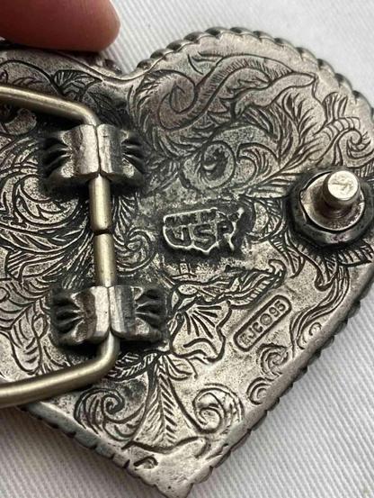 Back of a heart-shaped, ornate belt buckle with detailed floral engravings and a metal clasp.