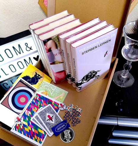 Foto von Fees Romanen »Sterben lernen« und »Die Schüchternheit der Pflaume« in einem Versandkarton, außerdem bunte Postkarten und zwei Fairy-Club-Buttons. Am Rand des Bildes ein Absinthglas.