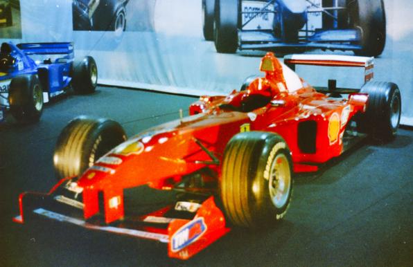 Photo d’une Ferrari F1 rouge éclatante, portant le numéro 3 de Michael Schumacher, exposée au Salon de l’Auto. Vestige des saisons 98-99-00, cette monoplace légendaire, issue de la collection du Prince Albert de Monaco, brille sous les projecteurs. Son aérodynamisme parfait, ses courbes taillées pour la vitesse et ses pneus lisses évoquent encore la fureur des circuits et la quête de gloire.