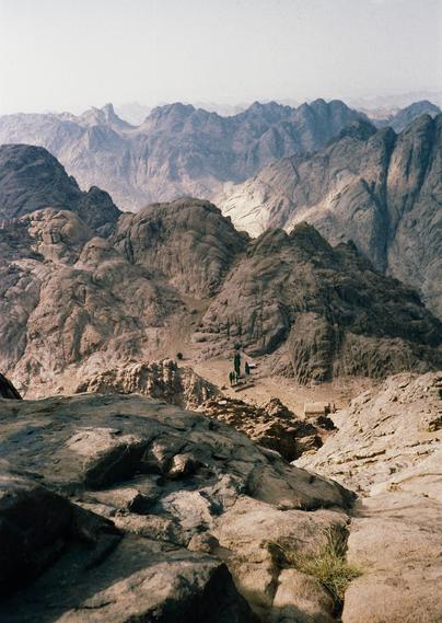Photo d' un paysage de montagne désertique, dans la péninsule du Sinaï en Égypte. Le terrain est rocheux et aride, avec des formations montagneuses aux formes distinctives. La couleur générale est un mélange de tons terreux, allant du beige clair au marron foncé, avec des nuances de gris et de bleu dans les zones plus éloignées.

Au premier plan, on voit un gros rocher avec une surface texturée, éclairée par la lumière du jour. À l'arrière-plan, plusieurs chaînes de montagnes s'étendent sur l'horizon, créant une perspective de profondeur. Les montagnes les plus proches sont plus détaillées et montrent des variations de couleurs et de textures, tandis que les montagnes plus éloignées s'estompent dans un bleu brumeux, suggérant une distance importante.
 
