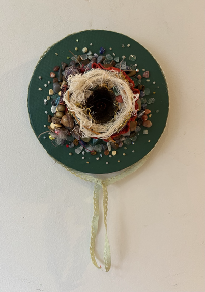 Small bird’s nest mounted on a round, green stretched canvas. Small coloured sones are glued to the canvas around the nest very close together at the bast and less close as the stones radiate outwards.