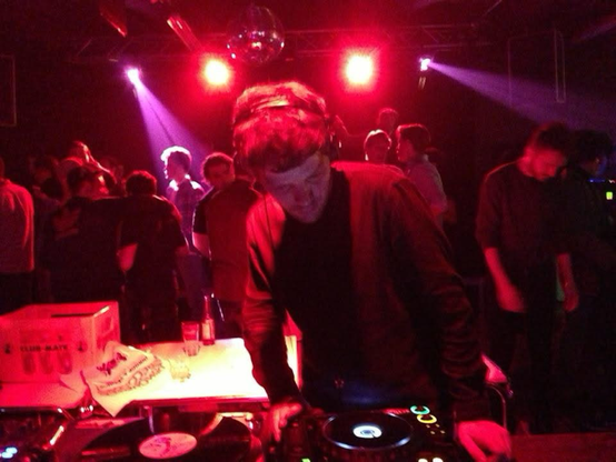 Red lighted photo of a DJ at the turntables. Concentrated. people in the back dancing.