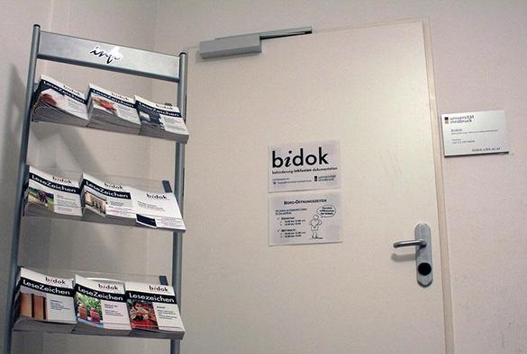 Büro-Eingang von bidok in der Sillgasse 8 im ersten Stock. Neben der Eingangs-Türe steht ein Broschüren-Ständer mit den zuletzt veröffentlichten "bidok LeseZeichen" zur freien Entnahme.