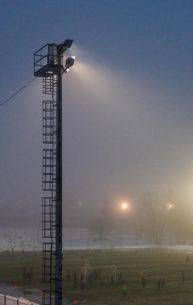Floodlights are trying to break through the evening darkness and fog to illuminate the football match at the stadium.