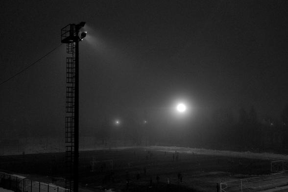 Floodlights are trying to break through the evening darkness and fog to illuminate the football match at the stadium.