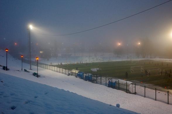 Floodlights are trying to break through the evening darkness and fog to illuminate the football match at the stadium.